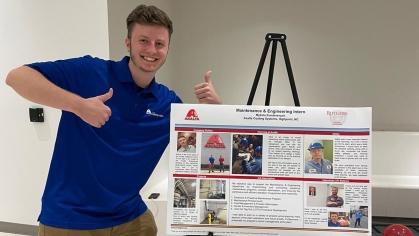 Young male student showing thumbs up next to a poster presentation on an easel.