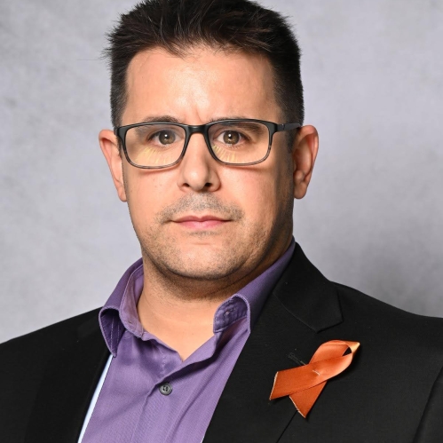 Head shot of white male with eyeglasses wearing a black suit jacket, a purple button down shirt and a breast cancer ribbon pinned to the lapel