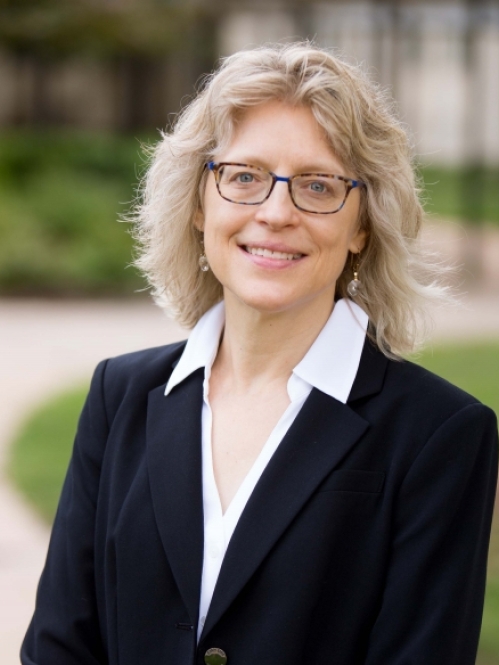 Woman with shoulder lenght blond hair and glasses wearing black suit with white blouse.