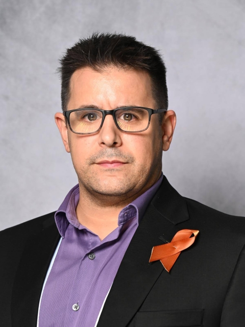Head shot of white male with eyeglasses wearing a black suit jacket, a purple button down shirt and a breast cancer ribbon pinned to the lapel