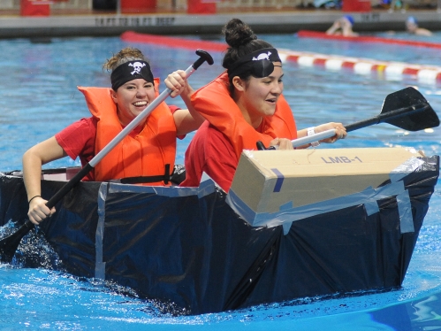 Two young women wearing orange life vests in a pool, paddling a boat made out of cardboard.