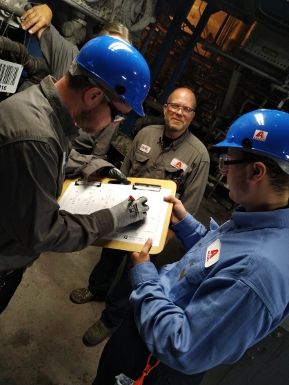 Two chemical plant workers wearing blue hard hats and safety goggles review charts on a clipboard asthird male looks on.