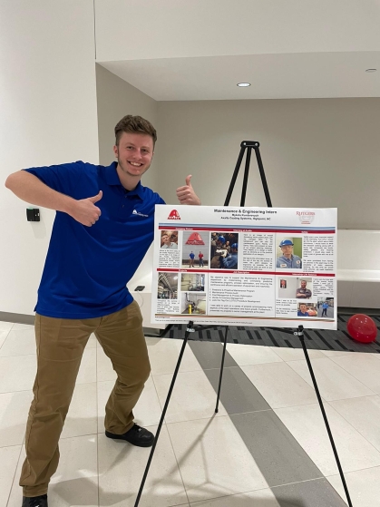 Young male student showing thumbs up next to a poster presentation on an easel.